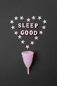 Pink menstrual cup on black background with with heart and words Sleep good. Concept possibility of use cup at night, zero waste, savings, minimalism. Feminine hygiene product, flat lay. Vertical.