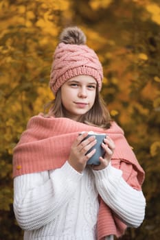 Portrait of pretty nice teenage girl drinking hot cocoa with marshmallows in autumn park