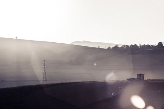 Sun appears over the hill to enlighten an electric tower and a small house in the middle of a field