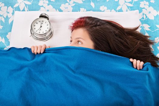 Portrait of a young woman lying in bed, hiding under a blue blanket and looking at the alarm clock. Student doesn’t want to wake up early for university or school. Oversleep, not enough sleep concept. Top view.