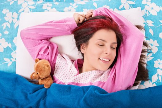Beautiful smiling young woman in pink pajamas sleeping in bed with a cute plush teddy bear.