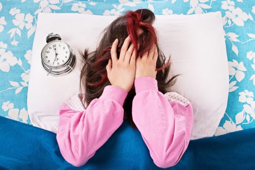 Young girl in pink pajamas lying in bed covering her face with her hands, annoyed and frustrated by the ringing of the alarm clock. Early morning concept.