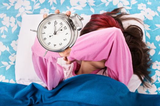 Young woman lying in bed covering her eyes with her arm, holding an alarm clock. Student doesn’t want to wake up early for university or school. Oversleep, not enough sleep concept. Top view.
