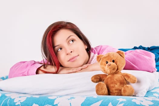 Beautiful young woman in pink pajamas lying in bed with her cute plush teddy bear, thinking of her dreams or a loved one.