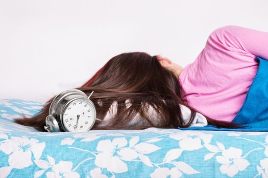 Beautiful young girl in pink pajamas calmly sleeping in her bed next to an old fashioned alarm clock, with her back turned to the camera. Healthy sleeping concept. Heavy sleeper concept.