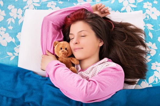 Beautiful young woman in pink pajamas sleeping in bed, hugging a cute plush teddy bear.