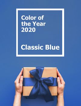 Female hands hold gift box on blue background with words Color of the Year 2020 Classic Blue. Gift box in craft wrapping paper with blue satin ribbon. Vertical.