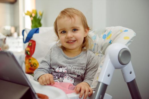 cute blond child sits in a children's chair at the kitchen and eat a juicy peach. Little girl with a piercing look.