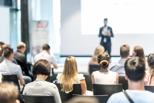 Business and entrepreneurship symposium. Speaker giving a talk at business meeting. Audience in conference hall. Rear view of unrecognized participant in audience.