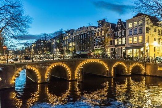 Amstedam, capital of Netherlands. Beautiful city of Amsterdam illuminated at dusk. Bicycles along the street and on the bridge over the canal.