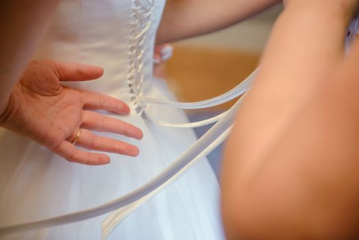 Bridesmaid helping slender bride lacing her wedding white dress, buttoning on delicate lace pattern with fluffy skirt on waist. Morning bridal preparation details newlyweds. Wedding day moments, wear.