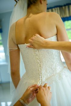 Bridesmaid helping slender bride lacing her wedding white dress, buttoning on delicate lace pattern with fluffy skirt on waist. Morning bridal preparation details newlyweds. Wedding day moments, wear.