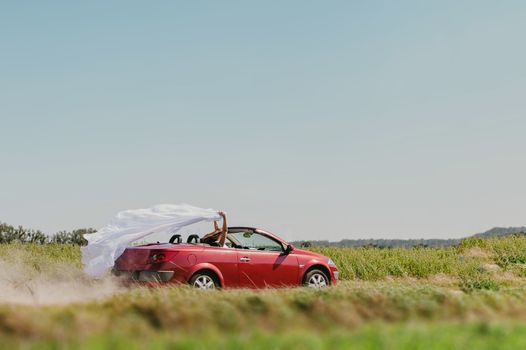 Bride and groom at a photo session in the nature. Moving fast red car with veil outside