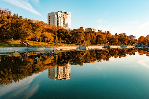 City ponds in the autumn. Trees covered with yellow and orange leaves are reflected in the water. Blue sky.