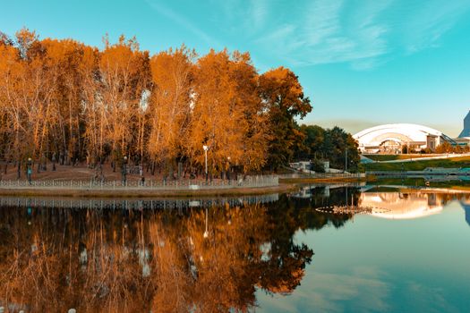 City ponds in the autumn. Trees covered with yellow and orange leaves are reflected in the water. Blue sky.