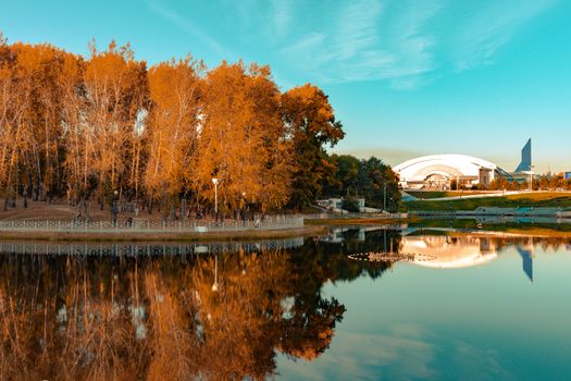 City ponds in the autumn. Trees covered with yellow and orange leaves are reflected in the water. Blue sky.