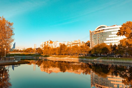 City ponds in the autumn. Trees covered with yellow and orange leaves are reflected in the water. Blue sky.