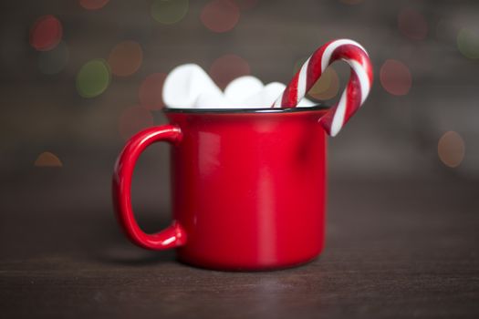 Cocoa in red mug with marshmallows and candy cane on dark wooden background with bokeh lights