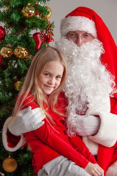 Portrait of smiling little girl in red clothes sitting on Santa Claus knees near christmas tree and smiling