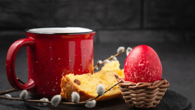 Red Egg in a wicker stand, Easter cake and cup of coffee on black slate dish and painted eggs on the table - traditional Easter breakfast.