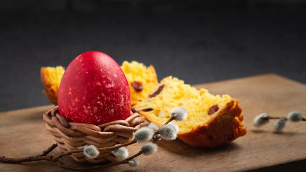 Red Easter egg in wisker stand and slice of Easter cake on a cutting board on a dark table - traditional Easter breakfast.