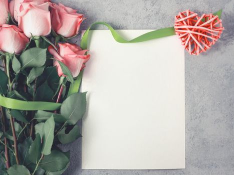 Flower arrangement - a bouquet of pink roses, a wicker heart and an empty sheet for inscription on a concrete surface, template for design or greeting card, top view, flat lay.