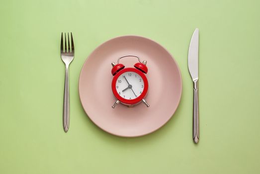 Red alarm clock on a clean pink plate flanked with knife and fork on a green background in a conceptual image