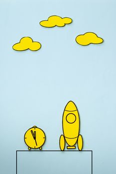 Little yellow rocket on a launch pad with alarm clock alongside and clouds above with copy space, cartoon illustration