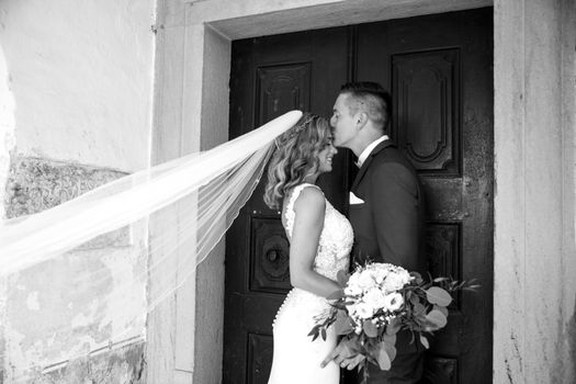 The kiss. Groom kisses bride on forehead in front of church portal. Close up portrait of sexy stylish wedding couple kissing. Black and white photo.