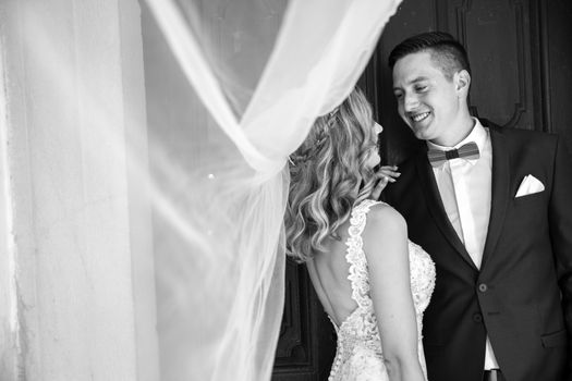 Groom hugs bride tenderly in the shadow of a flying veil. Close up portrait of sexy stylish wedding couple hugging under white vail. Artistic black and white wedding photo.