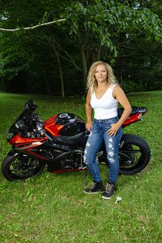 Twenty something blond woman, wearing a white tank to and jeans, standing in front of her motocycle