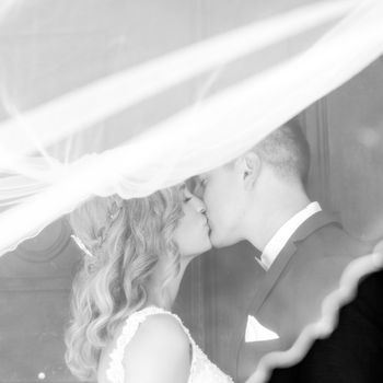 The Kiss. Bride and groom kisses tenderly in the shadow of a flying veil. Close up portrait of sexy stylish wedding couple kissing under white vail. Artistic black and white wedding photo.