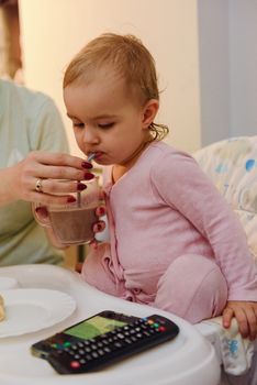 cute blond child sits in a children's chair at the kitchen and eat a juicy peach. Little girl with a piercing look.