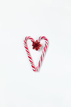 Christmas composition, heart made of two caramel candy canes and red bow on white background. Festive minimal style flat lay. For greeting card, invitation, social media. Vertical orientation.