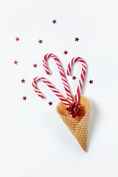 Christmas composition three caramel candy canes and bow in a waffle cone, confetti stars on white background. Festive minimal style flat lay. For greeting card, invitation. Vertical orientation.