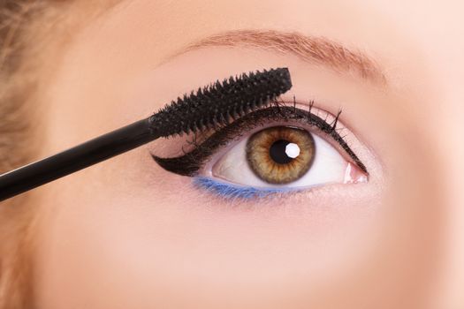 Close up of a female eye with cat eye black eyeliner and blue make-up, applying mascara with a brush on the eyelashes, isolated on white background. Close up shot of a female eye with clean make up.