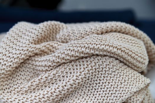 A soft knitted beige blanket made of natural wool lies rolled up against the background of a washed-out background. A close-up.