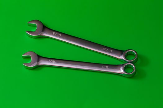 A pair of stainless steel wrenches, combination spanners with both an open end and a box end, lie on a solid green background. their sizes are engraved as fractions of an inch at 1/2" and 9/15".