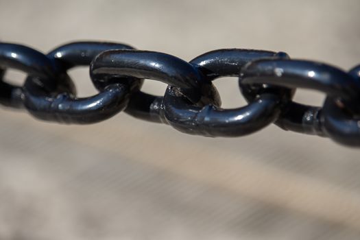 A close-up view in sunlight shows a metal utility chain painted in shiny and reflective black paint. The segment of chain is suspended above a blurry lighter background.