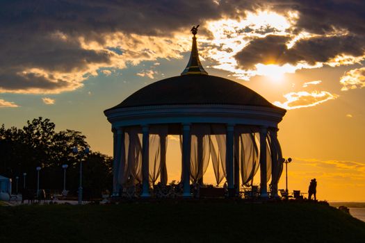 A couple in love hugs and kisses next to a romantic gazebo against the sunset