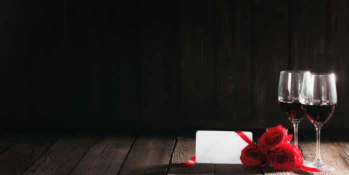 Two glasses of red wine, red roses and blank card on wooden background Valentines day concept