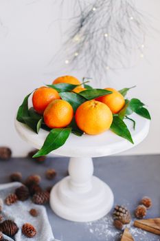 Orange tangerines on grey background in New Year's decor with brown pine cones and green leaves. Delicious sweet clementine. Christmas decoration with mandarins.