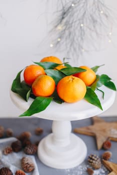 Orange tangerines on grey background in New Year's decor with brown pine cones and green leaves. Delicious sweet clementine. Christmas decoration with mandarins.