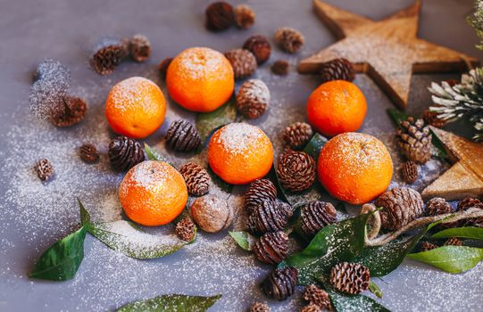 Orange tangerines on grey background in New Year's decor with brown pine cones and green leaves. Delicious sweet clementine. Christmas decoration with mandarins.