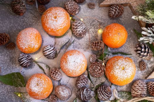 Orange tangerines on grey background in New Year's decor with brown pine cones and green leaves. Delicious sweet clementine. Christmas decoration with mandarins.