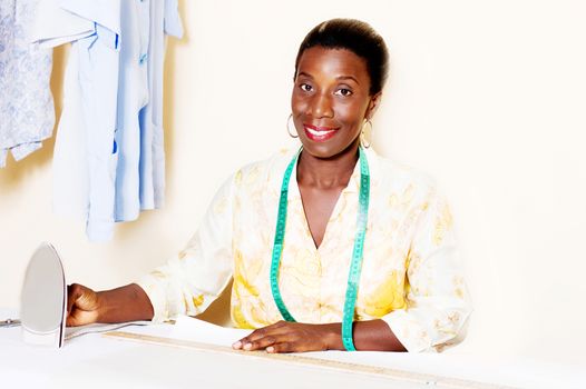 Young smiling seamstress holding an iron in front of her work table.