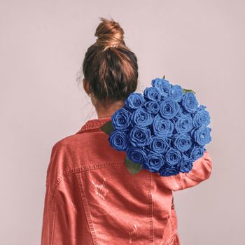 Back view of young woman in pink coral denim jacket holding bunch of blue roses on shoulder. Girl with bun updo in jeans holding flowers in blue color 2020. Copy space.