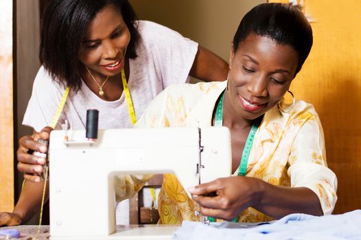 The professional seamstress sews and his trainee assists from behind.