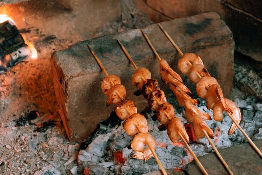 Shrimp frying on bamboo skewers on hot coals in a fireplace.