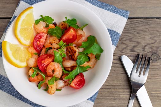 Fresh homemade salad of shrimp, arugula and tomato in a white plate on a wooden table, top view, flat lay, close up.